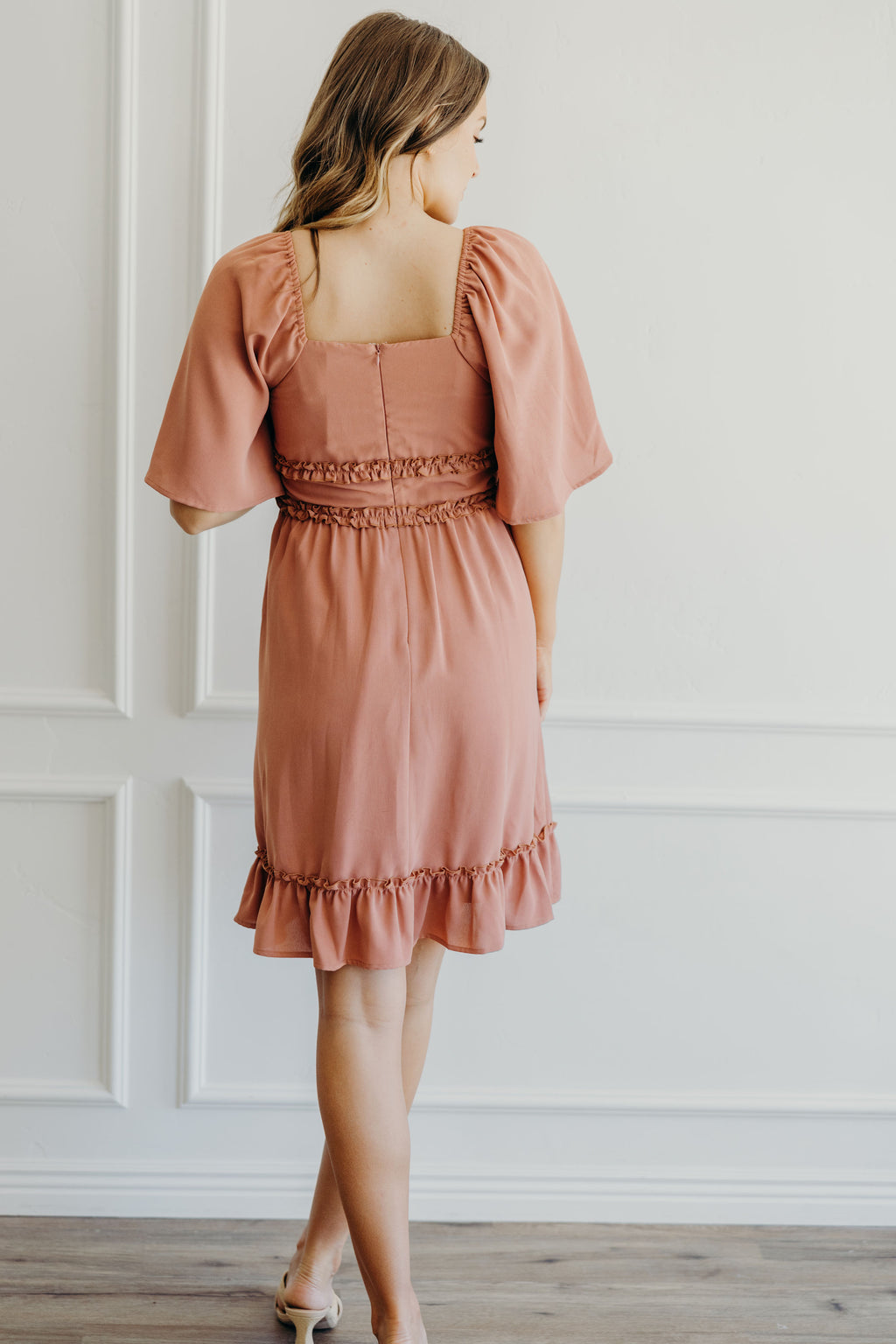 Back view of fit and flare pink dress with ruffle hem, ruffle detail at waist, back zipper, and oversized flutter sleeves.